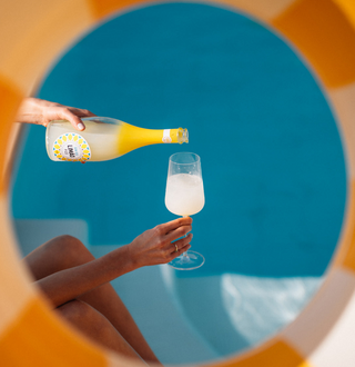 Gin Sul Limão Fizz wird am Pool ins Glas gegossen – Blick durch einen bunten Schwimmring auf Flasche und gefülltes Sektglas