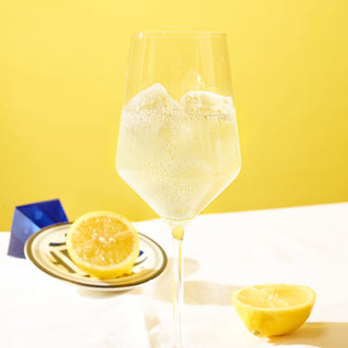 Weinglas mit Gin Sul Limão Fizz über Eis, halbierte Zitrone daneben, gelber Hintergrund